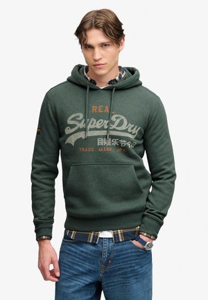 Ung mand med brunt hår iført en grøn SuperDry hættetrøje, ternet skjorte under, blå jeans og et ur med blå rem.