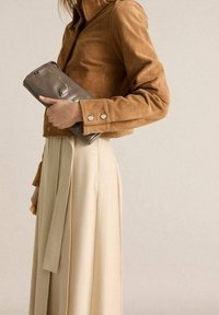 Bruine corduroy jas met zilverkleurige knopen, gecombineerd met een crèmekleurige rok en een kleine grijze clutch in de hand, die een strak ontwerp toont.