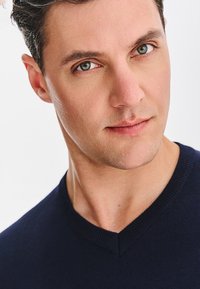 Granatowy sweter z dekoltem w serek, z prążkowanymi mankietami, gładką fakturą, dopasowany fason, noszony przez osobę o krótkich ciemnych włosach i jasnej karnacji.