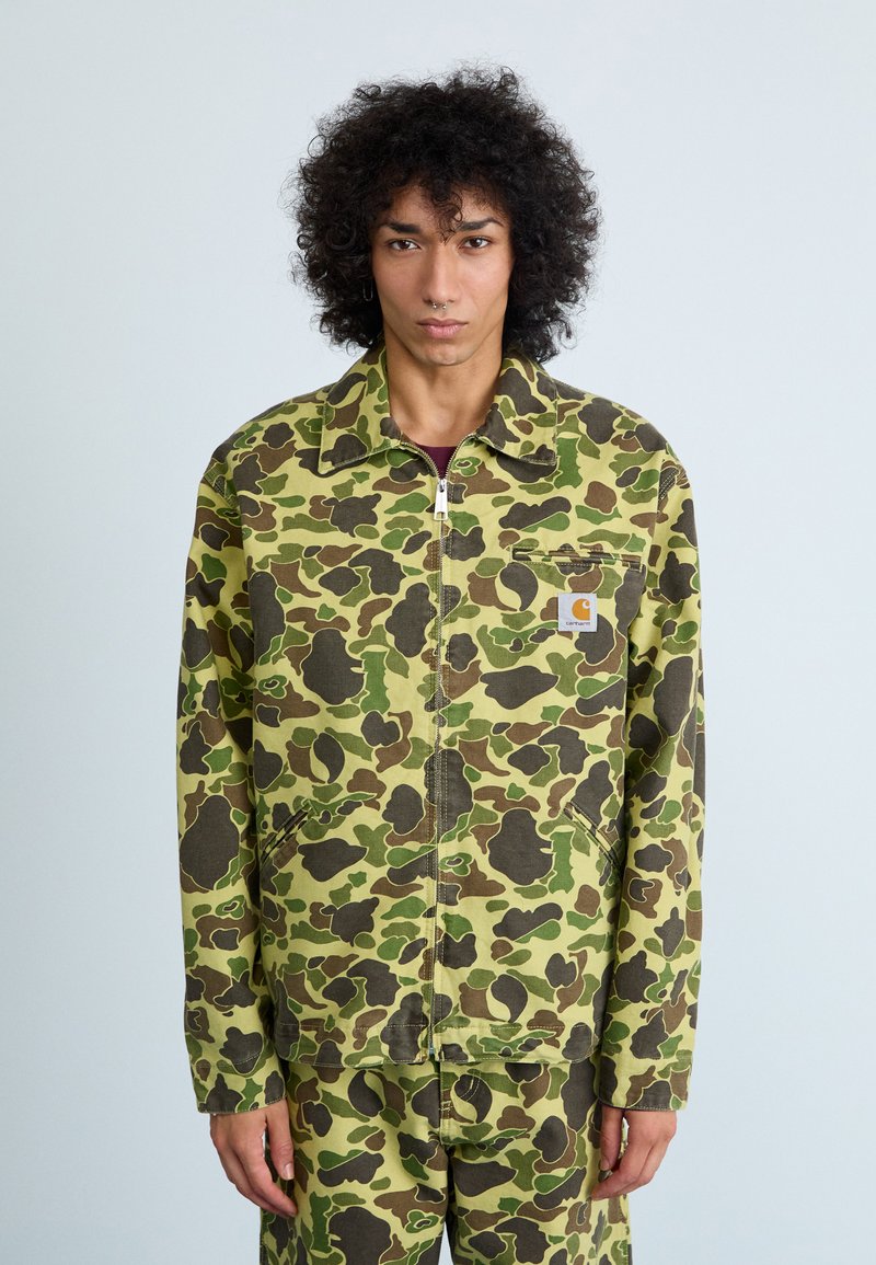 Jachetă cu fermoar în model camuflaj, în tonuri de verde, maro și negru; are guler și buzunare frontale; fabricată din bumbac cu o croială lejeră.