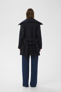 Tengerészkék kabát széles gallérral, derékban övvel és puha tapintású anyagból, világoskék bő szárú farmernadrággal párosítva. Hátulnézet.