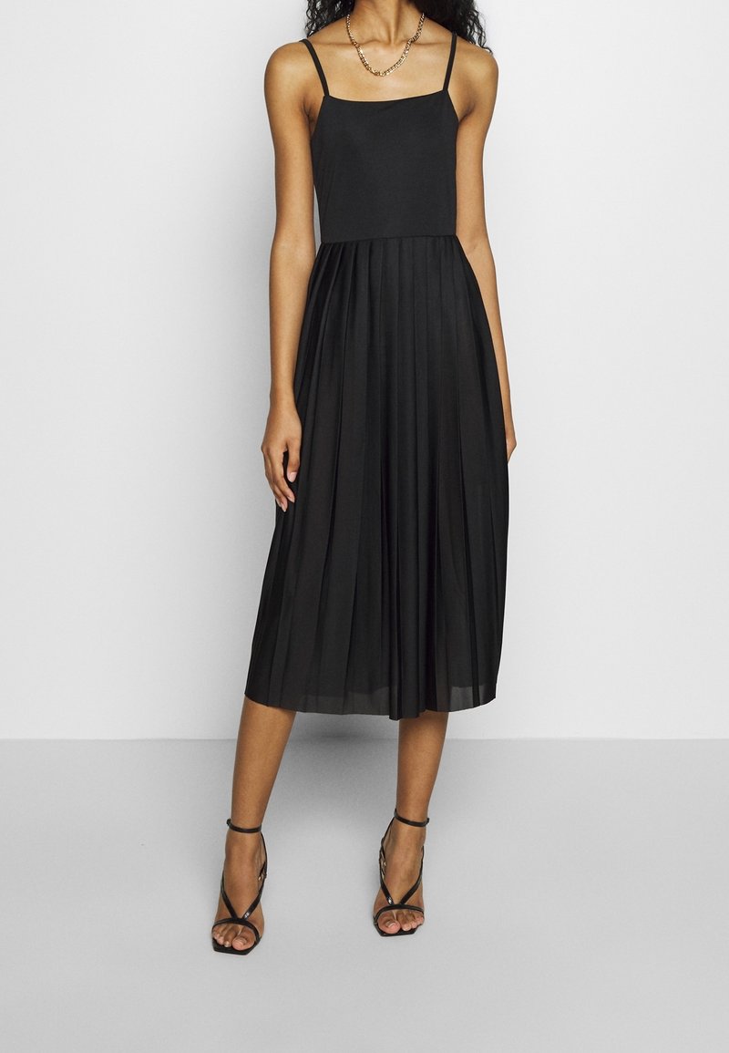 Femme portant une robe midi noire plissée à fines bretelles et des sandales noires à talons avec des lanières, debout devant un fond uni.