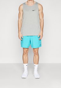 Camiseta sin mangas gris con logo negro, shorts turquesa y zapatillas blancas. El atuendo es minimalista, con una textura de tela suave.