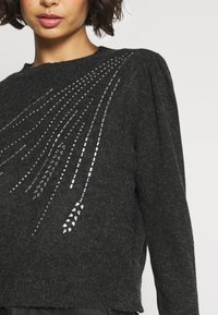 Ciemnoszary sweter o fakturowanym wykończeniu, ozdobiony srebrnymi cekinami ułożonymi w skośny wzór na przodzie i z bufiastymi rękawami.