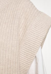 Beige gestrickte Sweaterweste mit breitem Rippenmuster und V-Ausschnitt. Der Stoff wirkt weich und leicht fusselig.
