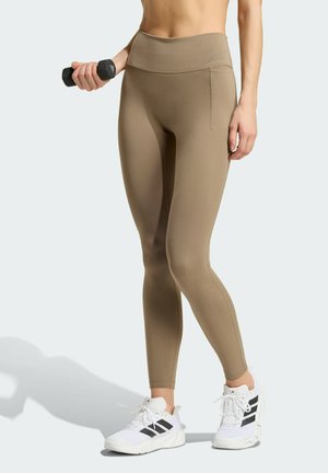 Femme portant des leggings beiges et des baskets blanches Adidas, tenant un haltère noir dans sa main droite.