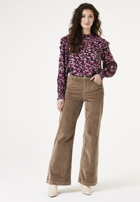 Langarmige, gerufflede blouse in donkerroze met abstract zwart-wit patroon. Hoge taille, wijde corduroy broek in taupe. Model draagt crèmekleurige laarzen.