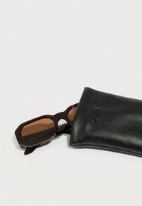 Očala za sonce z globoko bordo okvirjem in rjavimi lečami, ki počivajo delno znotraj črne usnjene zaščitne torbice z vtisnjenim napisom "QUAY".