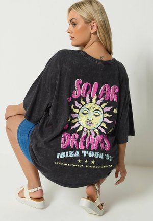 Blondynka kucająca, ubrana w czarny, oversize'owy T-shirt z kolorową grafiką słońca i napisem "Solar Dreams Ibiza Tour '97" na plecach.