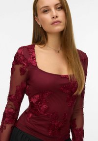 Ung kvinne med langt rett hår som har på seg en burgunder gjennomsiktig topp med blomsterbroderi, sølv øreringer i rund form, og et delikat anhengsmykke.