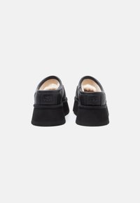 Par de sapatos slip-on UGG pretos com solas grossas e forro interior de cor creme, vistos de trás sobre um fundo branco.