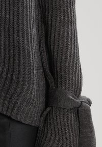 Pull en maille gris foncé montrant une texture côtelée, avec des manches longues et un détail de lien en tissu au niveau du poignet. Matière douce avec une légère apparence duveteuse.