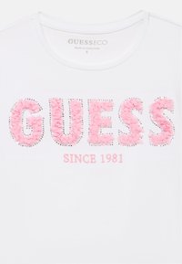Chemise blanche avec lettrage texturé rose "GUESS" bordé de petits clous argentés et "SINCE 1981" brodé en dessous en fil rose.