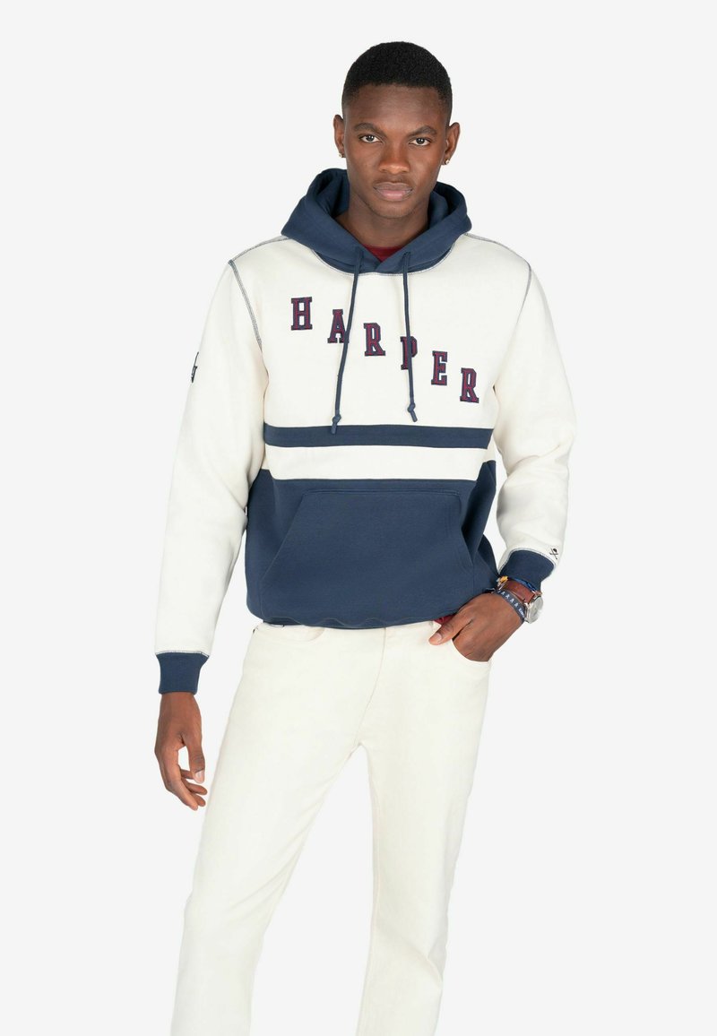 Hættetrøje i creme og navy, med "HARPER" i burgunderfarvede bogstaver, en frontlomme og ribbestrikkede manchetter. Båret med lyse bukser.