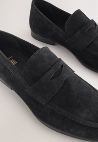 Mocassins en daim noir avec un bout arrondi, un petit talon et une bride décorative sur le dessus, sur un fond neutre.