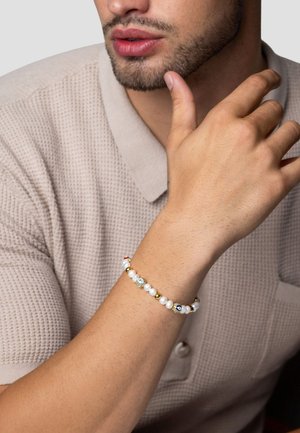 Perlesmykke med hvide perler, guld og farverige mønstrede accenter, sat mod en struktureret beige polo skjorte.