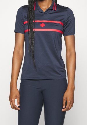 Donna che indossa una polo a maniche corte blu navy con strisce rosse e logo sul petto, abbinata a pantaloni blu navy.