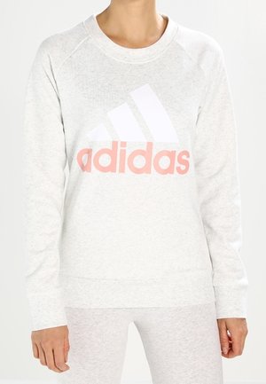 Femme portant un sweat-shirt Adidas gris clair avec un grand logo blanc et un texte rose à l'avant, assorti à un pantalon gris clair assorti.