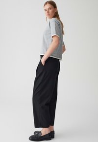 Grijs T-shirt met korte mouwen en witte afwerking, gecombineerd met zwarte losse broek. Model staat met de handen in de zakken en draagt zwarte instappers.
