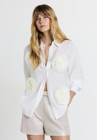 White button-up shirt with oversized white floral appliqués on pockets, made from a light, sheer fabric, paired with metallic silver shorts.