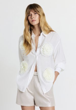 Weiße Bluse mit Knopfleiste und übergroßen weißen Blumenapplikationen auf den Taschen, aus leichtem, transparentem Stoff, kombiniert mit metallisch silbernen Shorts.