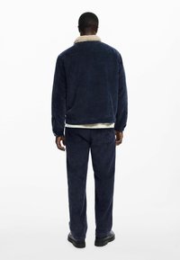 Tamno plavi kordura sako s kremastim sherpa ovratnikom, dugim rukavima i opuštenim krojem, uparen s odgovarajućim hlačama. Jednostavan dizajn, bez vidljivih dijelova.