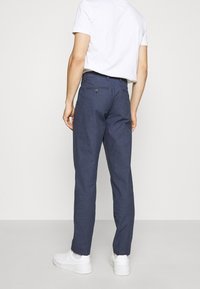 Pantalones ajustados azul marino hechos de una tela texturizada, con un bolsillo trasero con cierre de botón y un diseño ajustado, que se llevan con una camiseta blanca.