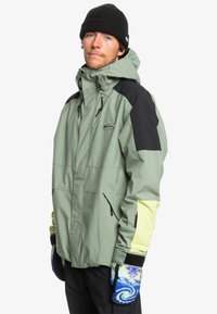 Lys grøn vandtæt jakke med sorte og gule accenter, flere lommer, justerbar hætte og mønstrede handsker. Slim fit.
