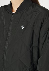 Czarna pikowana kurtka bomberka z zamkiem błyskawicznym z przodu, prążkowanym kołnierzem i małym białym logo na piersi. Gładki syntetyczny materiał.