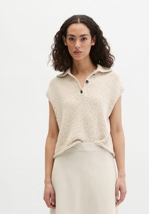 Kobieta o kręconych włosach, ubrana w bezrękawnik w kolorze beżowym wykonany na drutach z kołnierzykiem polo, włożony w wysokiej talii jasnobeżową spódnicę, na jednolitym tle.