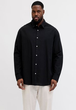 Homme portant une chemise noire à manches longues boutonnée et un pantalon clair, debout les mains détendues le long du corps.