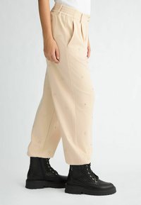 Beige Hosen aus weichem Stoff, mit einer lockeren Passform, Falten und dezenten gemusterten Verzierungen. Kombiniert mit schwarzen Schnürkampfstiefeln.