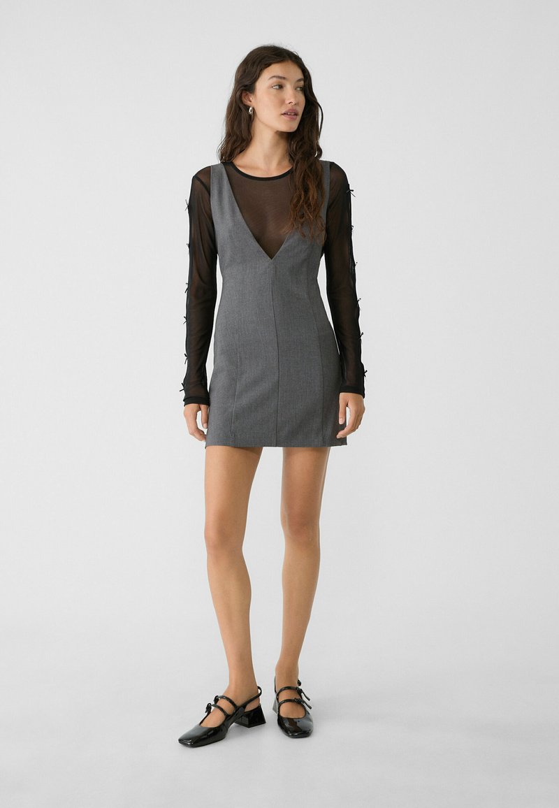 Abito grigio aderente con scollatura a V profonda, abbinato a un top trasparente nero a maniche lunghe con dettagli sottili sulle maniche.