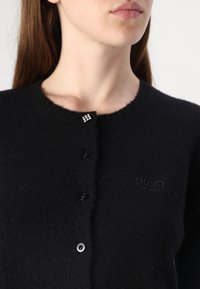 Cardigan nero lavorato a maglia con scollatura tonda, dotato di cinque bottoni neri e un accento di cristallo nel colletto. Materiale morbido e dalla texture piacevole.