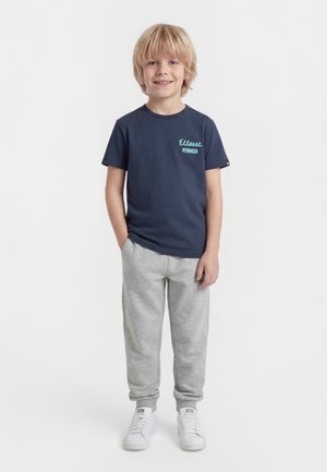 Νέος αγόρι που στέκεται, φοράει μπλε ναυτικό μπλουζάκι με ανοιχτό μπλε κείμενο, ανοιχτό γκρι φόρμας παντελόνι και λευκά αθλητικά παπούτσια, χαμογελαστός στην κάμερα.