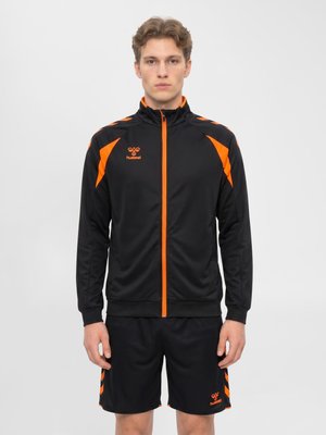 Młody mężczyzna w czarno-pomarańczowej sportowej kurtce i pasujących krótkich spodenkach stoi na jednolitym szarym tle.