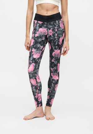 Femme portant des leggings noirs avec des motifs de roses roses et une ceinture noire, debout pieds nus sur un fond blanc uni.