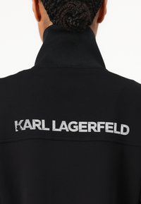 Čierna mikina s vysokým golierom, so strieborným logom "KARL LAGERFELD" na chrbte. Hladká textúra tkaniny, minimalistický dizajn.