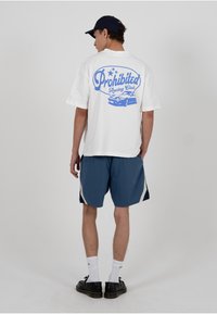 Valge lühikeste varrukatega t-särk, millel on sinine graafika tekstiga "Prohibited Racing Club" ja auto illustratsioon tagaküljel. Ühendatud siniste lühikestega.