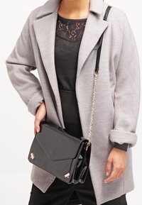 Black patent leather crossbody bag with a fold-over flap, gold accents, and a detachable chain strap. Smooth texture and structured design.