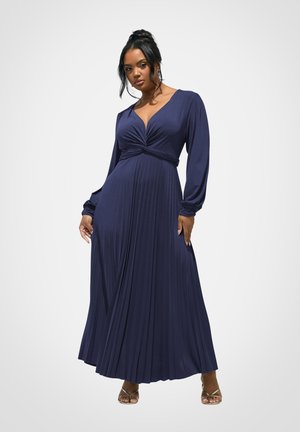 Kobieta stojąca w długiej granatowej plisowanej sukience z długimi rękawami i głębokim dekoltem w kształcie litery V, w połączeniu ze złotymi sandałami na obcasie z paskami.
