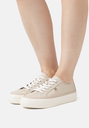 Jalat, jotka käyttävät beigejä matalia lenkkareita, joissa on valkoiset nauhat ja paksut valkoiset pohjat, yksinkertaista vaaleaa taustaa vasten.