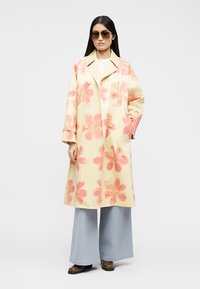 Γυναίκα που φοράει υπερμεγέθη κίτρινο παλτό με μεγάλα ροζ λουλούδια, φαρδιά ανοιχτό μπλε τζιν, καφέ παπούτσια και γυαλιά ηλίου, μπροστά σε απλό φόντο.