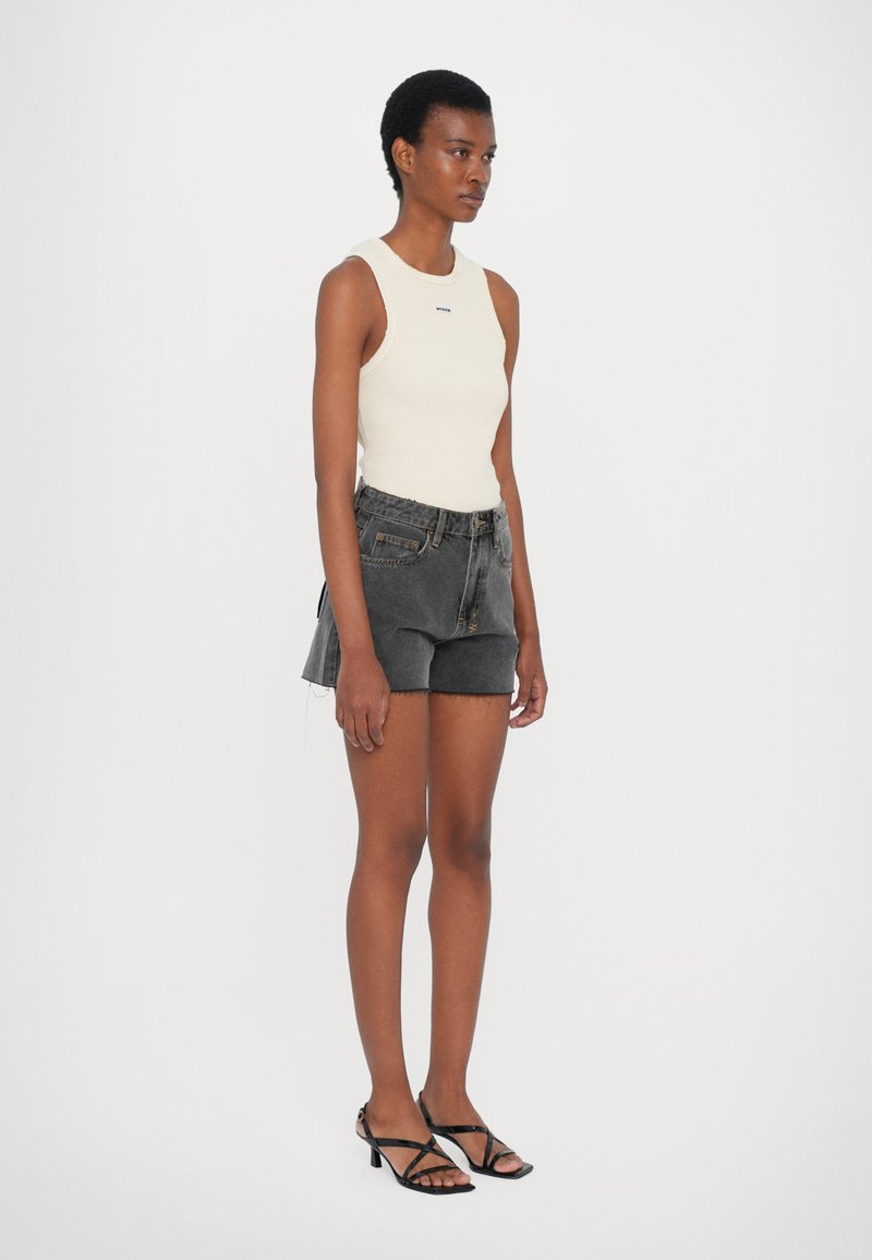 Top senza maniche crema, pantaloni shorts in denim neri con orlo sfilacciato e sandali neri con tacco. La modella è in piedi davanti a uno sfondo bianco uniforme.