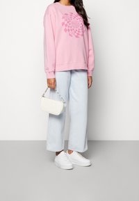 Rosa sweatshirt med grafiskt tryck, ljusblå vida jeans och vita sneakers. En liten vit axelväska med kedja hålls.