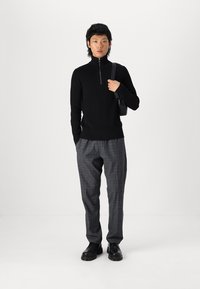 Un col roulé noir à côtes avec une fermeture éclair argentée, associé à un pantalon gris à carreaux et des chaussures noires, se tenant devant un fond blanc.