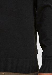 Gros plan sur une personne portant un pull noir, montrant l'ourlet et les poignets côtelés, avec une petite étiquette de marque visible sur le côté gauche.