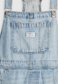 Salopette en denim bleu clair avec poche poitrine avant, boucles métalliques et une petite étiquette blanche Levi Strauss & Co sur la poche.