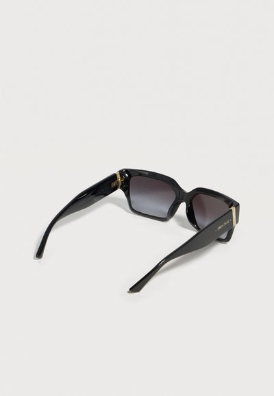 Μαύρα υπερμεγέθη τετράγωνα γυαλιά ηλίου με σκουρόχρωμους φακούς με βαθμιαία απόχρωση και χρυσές λεπτομέρειες στα μπράτσα, ακουμπισμένα ανοιχτά σε μια λευκή επιφάνεια.