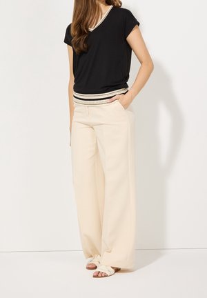 Mujer con top negro de manga corta con dobladillo a rayas, pantalones anchos color crema y sandalias blancas, de pie con una mano en el bolsillo.